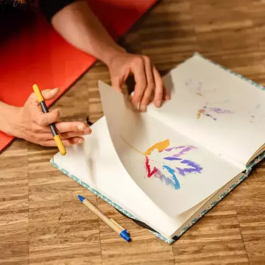 De armen van een persoon die in een schetsboek bladert. In het boek staat een geschilderd blad van een boom. De persoon houdt een gele stift in de hand. Er ligt een balpen naast het schetsboek. 